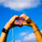 Hands form a heart shape around an American flag against a vibrant blue sky, symbolizing patriotism and freedom.