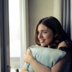 A woman sits comfortably by a window, holding a pillow and smiling happily indoors.