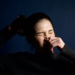 A woman in a dark sweater sneezes against a solid blue background, capturing a moment of candid expression.