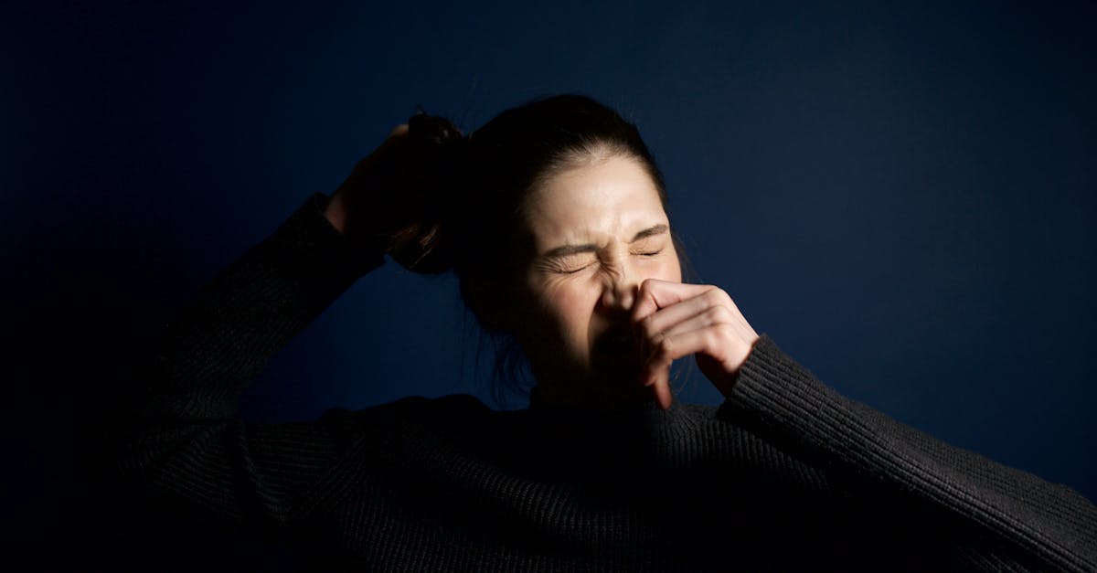 A woman in a dark sweater sneezes against a solid blue background, capturing a moment of candid expression.